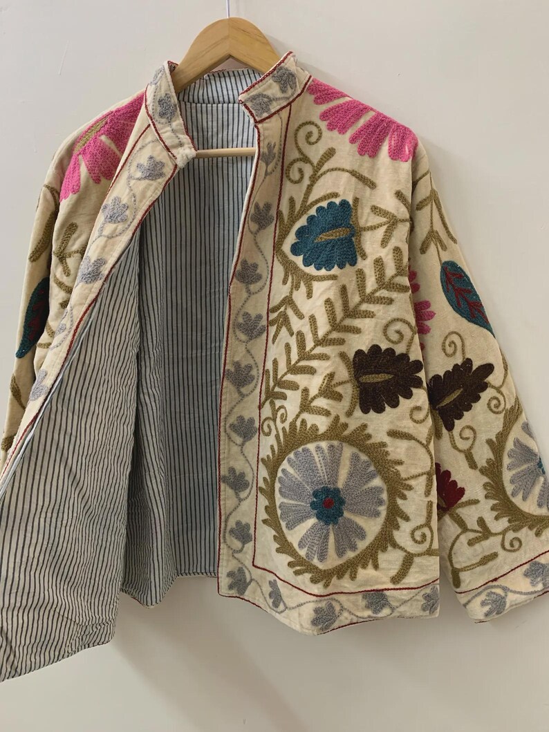 Puede incluir: Chaqueta beige bordada con dise&ntilde;os florales y de hojas en rosa, azul y marr&oacute;n. La chaqueta tiene un forro a rayas y ribetes rojos. La chaqueta est&aacute; colgada en una percha de madera.