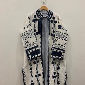 May include: A long, white coat with intricate dark blue floral embroidery and trim. The coat features a striped lining and is hanging on a wooden hanger. The design includes floral and geometric patterns.