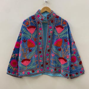 Puede incluir: Chaqueta bordada azul con cuello mandarín y diseño floral. La chaqueta presenta flores rosas y naranjas con hojas verdes. El forro interior es de tela a rayas azul claro y blanco.
