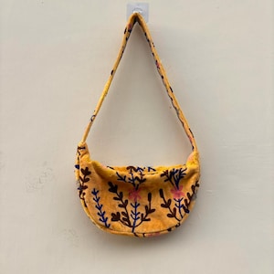 Pu&ograve; includere: Una borsa a tracolla a forma di mezzaluna realizzata in tessuto giallo dalla consistenza morbida. La borsa presenta un motivo ricamato di steli botanici e fiori marrone scuro, blu e rosa sul corpo e sull'unica tracolla integrata.