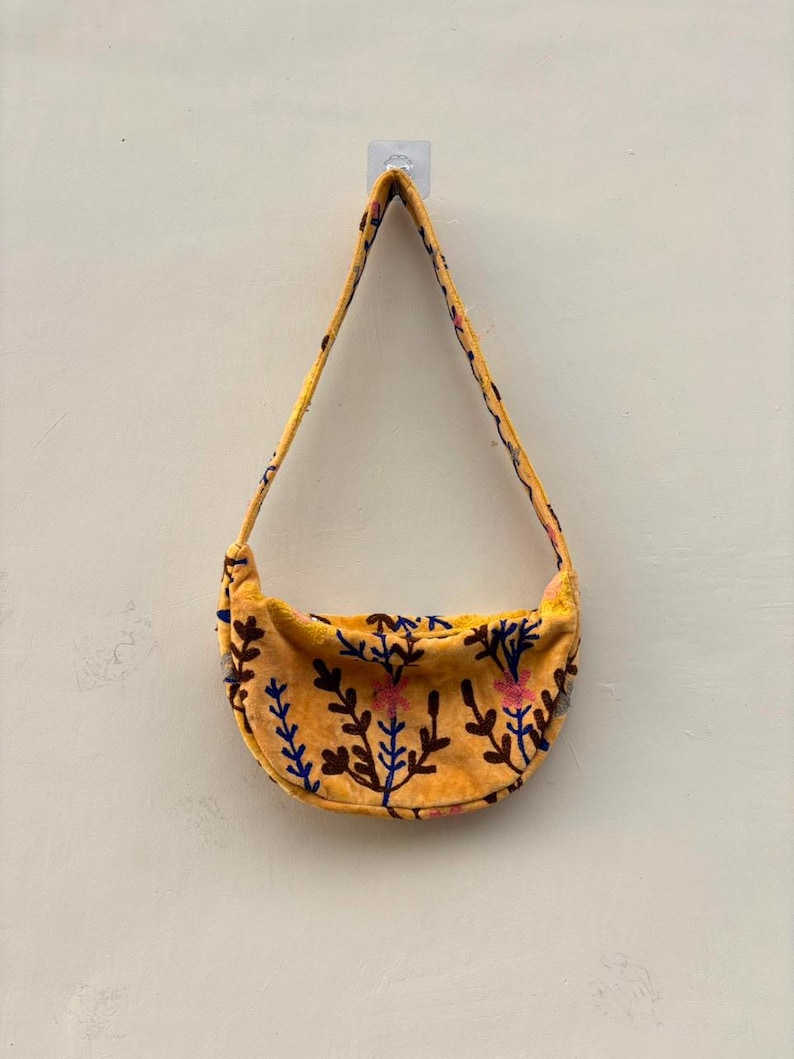 Pu&ograve; includere: Una borsa a tracolla a forma di mezzaluna realizzata in tessuto giallo dalla consistenza morbida. La borsa presenta un motivo ricamato di steli botanici e fiori marrone scuro, blu e rosa sul corpo e sull'unica tracolla integrata.