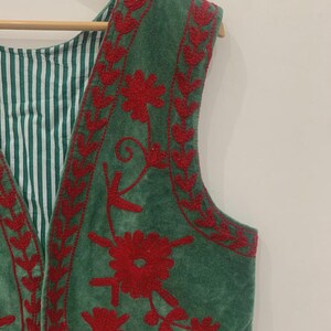 K&ouml;nnte beinhalten: Eine gr&uuml;ne Weste mit roter Blumenstickerei. Die Weste hat ein gestreiftes gr&uuml;n-wei&szlig;es Futter und h&auml;ngt an einem Holzb&uuml;gel. Die Stickerei ist detailliert und bedeckt die R&auml;nder und die Vorderseite der Weste.