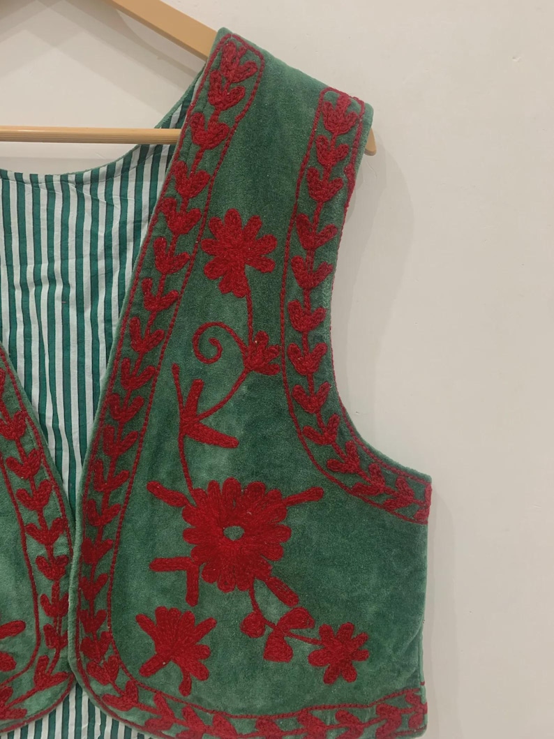 K&ouml;nnte beinhalten: Eine gr&uuml;ne Weste mit roter Blumenstickerei. Die Weste hat ein gestreiftes gr&uuml;n-wei&szlig;es Futter und h&auml;ngt an einem Holzb&uuml;gel. Die Stickerei ist detailliert und bedeckt die R&auml;nder und die Vorderseite der Weste.