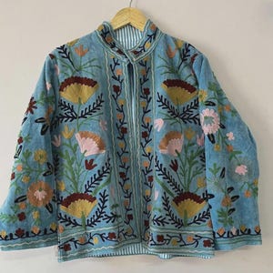 May include: A light blue jacket with intricate floral embroidery in shades of yellow, brown, pink, and green. The jacket features a mandarin collar and a striped lining. The embroidery is detailed and covers the front and sleeves.