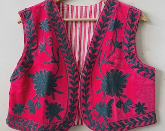 May include: A hot pink vest with dark teal floral embroidery and leaf trim. The vest's interior lining features a red and white striped pattern. The vest is sleeveless and hangs on a wooden hanger.