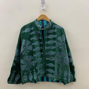 Könnte beinhalten: Dunkelgrüne Samtjacke mit gestickten Blumen- und abstrakten Designs. Die Jacke hat einen Stehkragen und ist mit einem blau-grün gestreiften Stoff gefüttert. Sie hängt an einem Holzbügel.