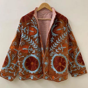 以下が含まれることがあります： オレンジと水色の花柄刺繍が施された茶色のジャケット。 ジャケットにはストライプの裏地が付いており、木製のハンガーに掛けられています。 刺繍はジャケットの表面に詳細なパターンを作り出しています。
