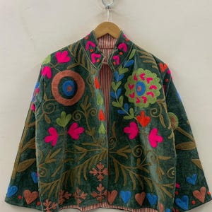 May include: A dark green velvet jacket with colourful floral embroidery. The jacket features bright pink, orange, and blue flower and heart designs, with brown and gold accents. The interior lining has a red and white striped pattern.
