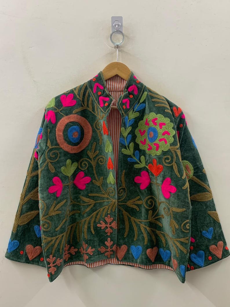 May include: A dark green velvet jacket with colourful floral embroidery. The jacket features bright pink, orange, and blue flower and heart designs, with brown and gold accents. The interior lining has a red and white striped pattern.