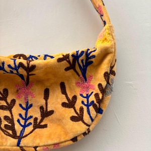Pu&ograve; includere: Una borsa a tracolla gialla a forma di mezzaluna con un motivo floreale ricamato composto da steli marroni, foglie blu e fiori rosa. La borsa presenta una tracolla gialla abbinata e una superficie morbida e testurizzata con ricami.