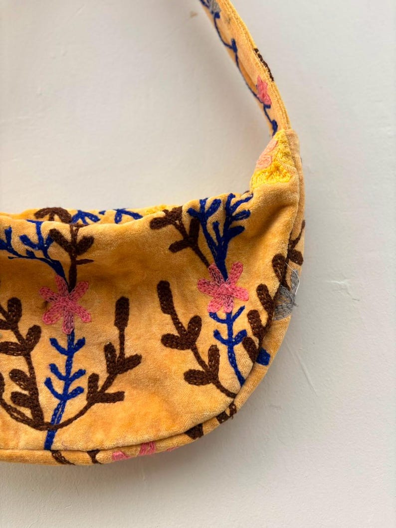 Pu&ograve; includere: Una borsa a tracolla gialla a forma di mezzaluna con un motivo floreale ricamato composto da steli marroni, foglie blu e fiori rosa. La borsa presenta una tracolla gialla abbinata e una superficie morbida e testurizzata con ricami.