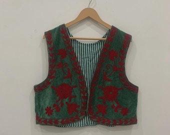 May include: Green velvet vest with red floral embroidery. The vest has a striped lining and is sleeveless. It is hanging on a wooden hanger against a white wall.
