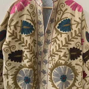 Puede incluir: Chaqueta bordada de color crema con patrones florales en rosa, turquesa, marr&oacute;n y gris. La chaqueta tiene cuello mandar&iacute;n y forro a rayas. El bordado presenta intrincados dise&ntilde;os florales y de hojas, creando una est&eacute;tica bohemia.