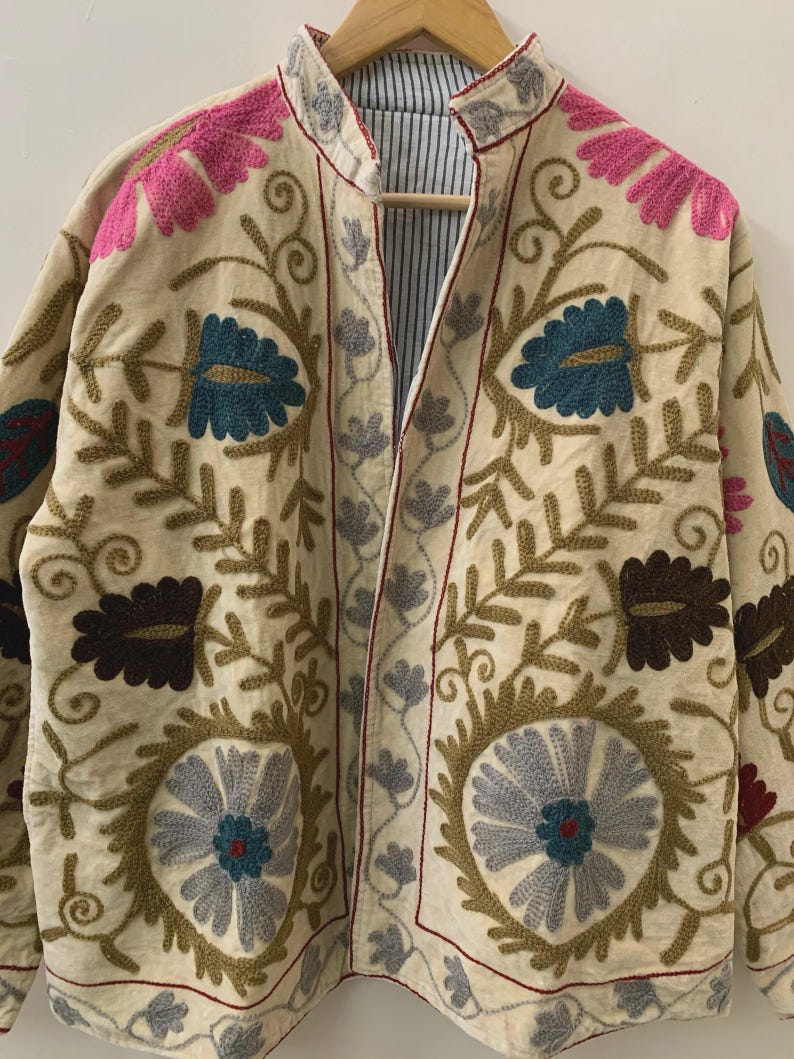 Puede incluir: Chaqueta bordada de color crema con patrones florales en rosa, turquesa, marr&oacute;n y gris. La chaqueta tiene cuello mandar&iacute;n y forro a rayas. El bordado presenta intrincados dise&ntilde;os florales y de hojas, creando una est&eacute;tica bohemia.