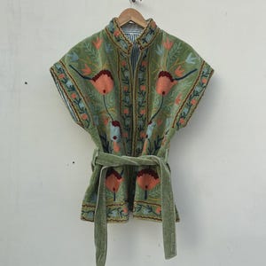 Op de afbeelding: Een saliegroene geborduurde jas met een opstaande kraag en korte mouwen. De jas heeft een bloemenmotief in tinten oranje, blauw en bruin. Hij heeft een bijpassende riem en hangt aan een houten hanger.