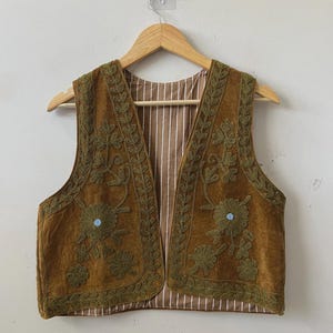 May include: A brown vest with olive green floral embroidery. The vest has a striped brown and white lining. The embroidery includes floral and leaf patterns. The vest is hanging on a wooden hanger.