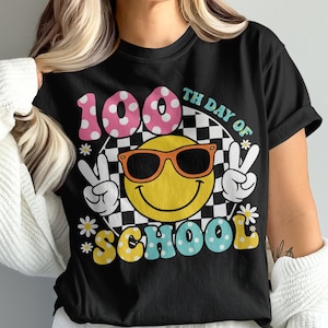 May include: Black t-shirt with a colorful graphic for the 100th day of school. The design features a smiley face wearing sunglasses, peace signs, and the words "100th Day of School". The number "100" is in pink polka dots.