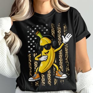 May include: Black t-shirt featuring a cartoon banana wearing sunglasses and headphones, dabbing. The background has an American flag design with leopard print stripes. The banana is wearing orange sneakers.