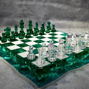 May include: A chess set featuring a square board with a translucent emerald green, textured, uneven edge and a grid of alternating white and green squares. The set includes clear, colorless glass pieces and solid emerald green glass pieces.