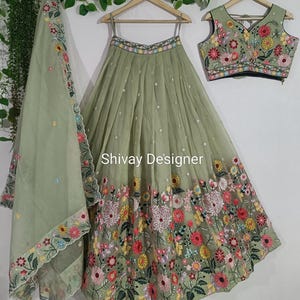 Puede incluir: Un conjunto lehenga bordado en verde salvia con un dupatta y una blusa a juego. El lehenga presenta un borde bordado floral con un escote en V y tirantes finos. El texto "Shivay Designer" es visible en la falda.