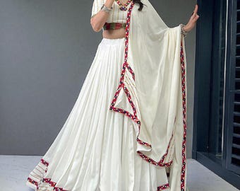 May include: A white, pleated skirt and matching top with a red and gold embroidered border. The outfit includes a white shawl with a similar border design. The top has a floral pattern on the midriff.