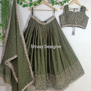 Puede incluir: Conjunto de ropa étnica de tres piezas verde oliva. El conjunto incluye una falda plisada, un top sin mangas y una bufanda a juego. La falda y la bufanda tienen intrincados bordados dorados en los bordes. El texto "Shivay Designer" es visible en la falda.