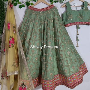 Puede incluir: Falda y top bordados en verde salvia, con una bufanda amarilla con estampado floral. La falda y el top están adornados con bordados dorados. El texto "Shivay Designer" es visible en la falda.
