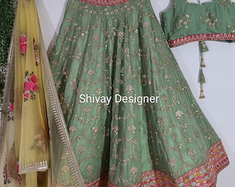 May include: Sage green embroidered skirt and matching top with a yellow floral patterned scarf. The skirt and top are embellished with gold-colored embroidery. The text "Shivay Designer" is visible on the skirt.