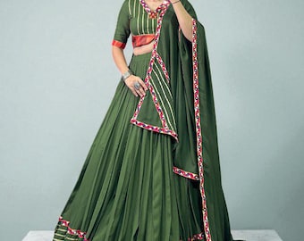 May include: An olive green traditional Indian outfit, featuring a long pleated skirt, a cropped top, and a matching scarf. The outfit is adorned with gold, red, and white trim. The top has short sleeves and a V-neckline. The outfit is designed with a traditional Indian aesthetic.