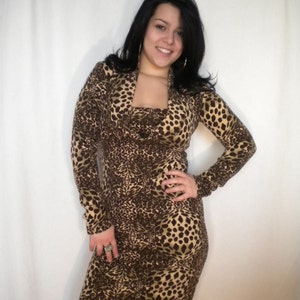 May include: A long-sleeved, body-hugging dress with a cowl neckline. The dress features a brown and tan leopard print pattern. The model is wearing a ring and earrings. The dress is knee-length.
