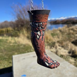 Könnte beinhalten: Ein keramischer, beinartiger Hängeornament in Dunkelrot und Schwarz. Der Fuß hat weiße Akzente. Es wird von einer silbernen Kette gehalten. Der Hintergrund zeigt eine natürliche Umgebung mit blauem Himmel und See.