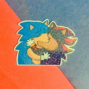 May include: A holographic sticker featuring a blue hedgehog and a black hedgehog embracing. The blue hedgehog has tan accents and white gloves, while the black hedgehog has red markings and white gloves. Both characters have closed eyes and a glittery finish.