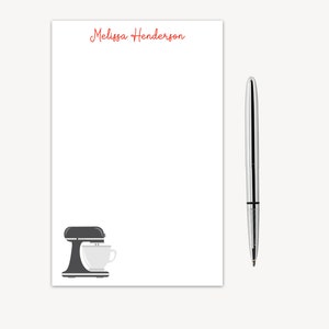May include: A white notepad with the text "Melissa Henderson" at the top. A black and white illustration of a stand mixer is at the bottom. A silver pen is next to the notepad.