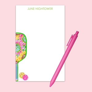 May include: A white notepad with the name "JUNE HIGHTOWER" printed at the top. A colorful floral patterned paddleball racket is on the left side of the notepad. Two small colorful balls are below the racket. A pink pen is lying on the notepad.