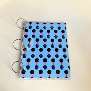 May include: A blue and white patterned notebook with a clear plastic cover and three silver rings. The cover features a repeating pattern of small green topiary trees in black pots with pink bows.