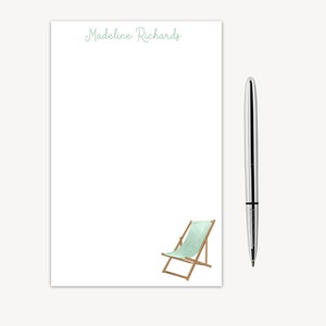 May include: A white notepad with the name "Madeline Richards" printed at the top. A light blue and brown deck chair is illustrated at the bottom of the notepad. A silver pen is next to the notepad.