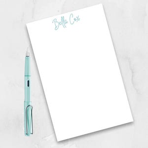 May include: A white notepad with the name "Bella Cox" written in blue cursive at the top. A light blue fountain pen is lying on a white surface next to the notepad.