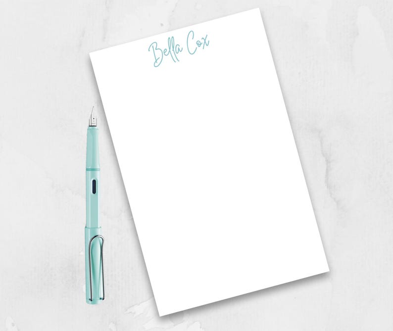 May include: A white notepad with the name "Bella Cox" written in blue cursive at the top. A light blue fountain pen is lying on a white surface next to the notepad.