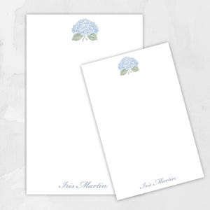 May include: Two white notepads with a light blue hydrangea flower illustration at the top. The name "Iris Martin" is printed in a light blue script at the bottom of each notepad. One notepad is larger than the other.