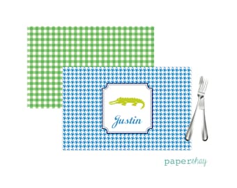 May include: Personalized placemat with a green alligator design and the name "Justin" in a blue and white houndstooth pattern. The placemat is green and white gingham.