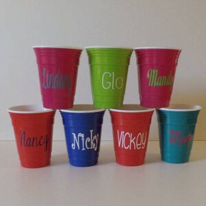 Op de afbeelding: Een verzameling kleurrijke plastic bekers, elk met een witte rand en een gepersonaliseerde naam in een sierlijk lettertype. De bekers zijn in tinten rood, groen, blauw en turquoise. De namen zijn "Lindsey", "Glo", "Mandy", "Nancy", "Nicky", "Vickey" en "Mary".