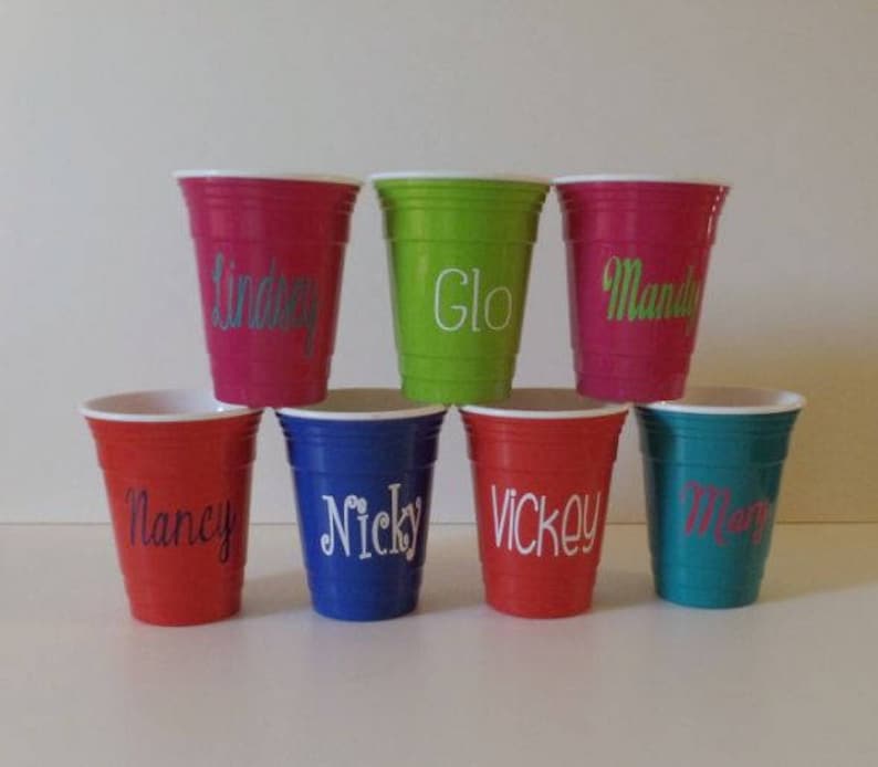 Op de afbeelding: Een verzameling kleurrijke plastic bekers, elk met een witte rand en een gepersonaliseerde naam in een sierlijk lettertype. De bekers zijn in tinten rood, groen, blauw en turquoise. De namen zijn "Lindsey", "Glo", "Mandy", "Nancy", "Nicky", "Vickey" en "Mary".