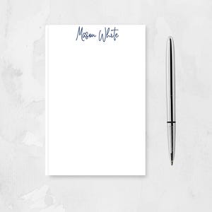 May include: A white notepad with the name "Mason White" written in blue ink on the top. A silver pen is next to the notepad.