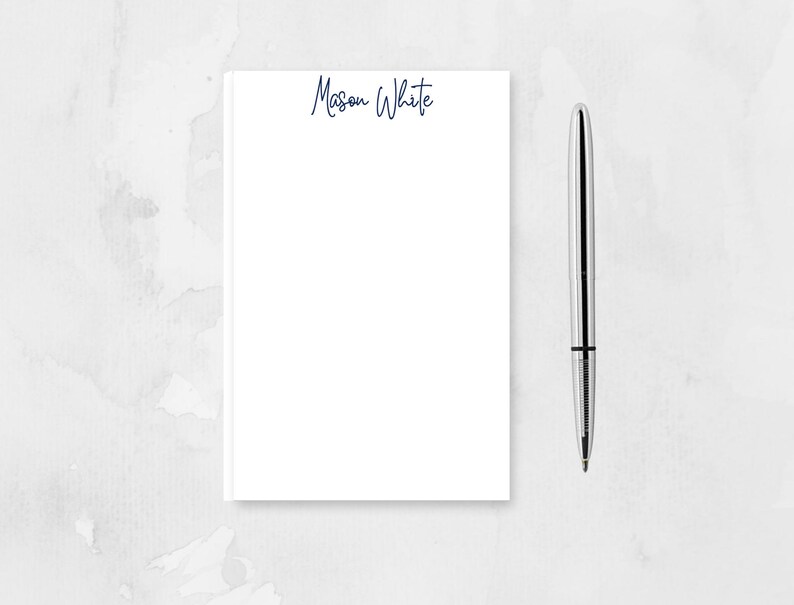 May include: A white notepad with the name "Mason White" written in blue ink on the top. A silver pen is next to the notepad.