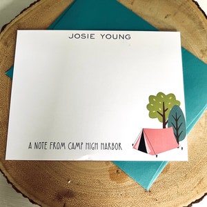 May include: A white card with the text "JOSIE YOUNG" at the top and "A NOTE FROM CAMP HIGH HARBOR" at the bottom. A pink tent with a black flap is illustrated in the bottom right corner of the card, with two green trees behind it.