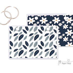 May include: Two clear plastic sheets with different floral patterns. The top sheet has a navy blue background with white daisies. The bottom sheet has a white background with navy blue and gray leaves. Both sheets have silver rings in the top corners.