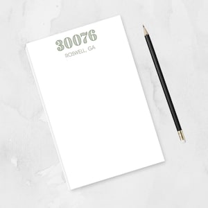 May include: A white notepad with the text "30076 Roswell, GA" printed in a light green color. A black pencil with a silver tip rests on the right side of the notepad. The background is a light gray watercolor wash.