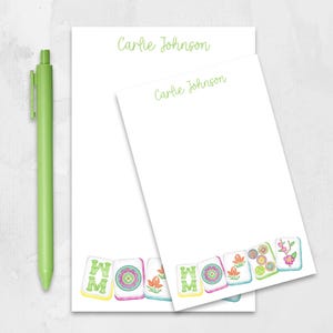 May include: Two white notepads with the name "Carlie Johnson" in green script. The bottom of the notepads feature colorful watercolor illustrations of Mahjong tiles. A green pen is positioned to the left of the notepads.
