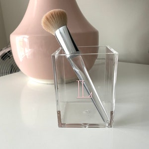May include: A clear acrylic makeup brush holder with a pink "II" monogram. A silver makeup brush is inside the holder.