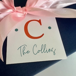 May include: A white gift tag with a large red letter "C" and the text "The Collins" in teal green script. The tag is tied with a pink satin ribbon.